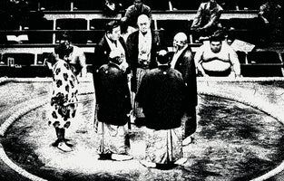 A grainy, high-contrast, black-and-white photograph of a sumo wrestling ring from a high angle. Several officials in traditional Japanese robes are gathered in the center of the ring, known as a dohyō, appearing to be in a discussion. Other wrestlers are seated around the ring in the background.