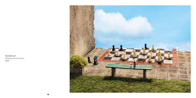 A page layout showing an artwork titled Spielabbruch, Breaking Off the Game, 2019. The art is a surreal photograph of a large chessboard on a stone patio against a cloudy blue sky. A chess game is in progress, but a black knight and pawn are standing off the board. In the foreground, a green bench holds a bottle of wine and a glass.
