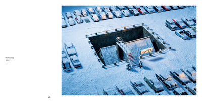 A high-angle view of a miniature diorama depicting a snow-covered parking lot filled with vintage cars. In the center, a large, rectangular pit is carved out, revealing a small, submerged building. A narrow staircase leads down to the structure, which has a small, illuminated sign that reads HOTEL.