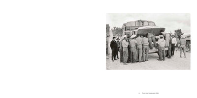 A black and white photograph from 1964 shows a group of men in Guatemala gathered around the open hood of a large truck bus on a dirt road. Several men are bent over, looking into the engine compartment, while others stand by and watch, suggesting the vehicle has broken down. The truck is heavily loaded with large bundles on its roof.