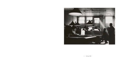 A moody, black and white photograph of a pool hall from 1956. Numerous conical lamps hang low over several billiard tables. In the background, the silhouettes of two men stand near a window as a dramatic shaft of sunlight cuts through the hazy, dim room.