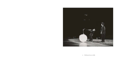 A black and white photograph of a PNE Moscow Circus performance, circa 1963. On a dimly lit stage, one performer in a uniform leans back to play a clarinet upwards. Another performer stands next to him, watching. A large bass drum is positioned on the stage beside them.
