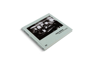 A square hardcover book with a pale green cloth cover, titled Fred Herzog Black and White. The book is set against a white background and features a high-contrast, black and white photograph of an interior scene on the front.
