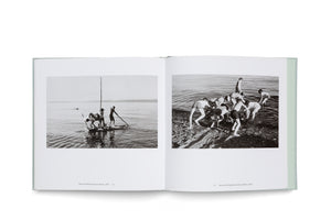 An open photobook with a pale green cover is laid flat against a white background. The two-page spread features black-and-white photographs from 1970. On the left page, three boys are on a homemade raft on the water. On the right page, a group of boys on a rocky shore work together to launch a raft.