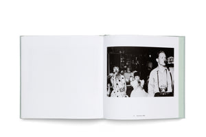An open book with a pale green cover lies on a white surface. The left page is blank, while the right page features a black-and-white photograph captioned Soda Shop, 1958. The photo shows a woman, a young girl, and a man in a diner, all drinking from glass bottles.