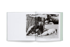An open book with a light green cover displays a black-and-white photograph on the right page. The photo shows two people on a sandy beach next to a large tree stump. One person sits and looks through binoculars, while the other lies on their back, sunbathing.