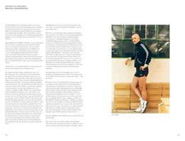 A book spread with German text on the left and a color photograph on the right. The photo is a full-length portrait of a man with a shaved head and a mustache, wearing a black Adidas tracksuit top, black shorts, and white sneakers. He leans against a white wall next to a window, resting one arm on a stack of cardboard boxes.