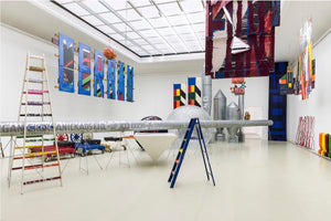 A wide-angle interior view of a contemporary art exhibition in a large, white gallery. The space is filled with a diverse installation of colorful, abstract objects, including a long silver tube on stands, painted panels hanging from the ceiling, several step ladders, and silver silo-shaped sculptures in the background.