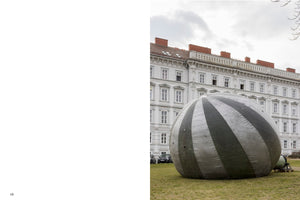 A book page with a photograph on the right showing a giant, inflatable sphere with wide, alternating silver and dark grey stripes. The object sits on a grassy lawn in front of a large, ornate, white classical building under an overcast sky.