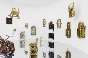 A wide-angle view of a contemporary art installation in a white, curved gallery with a highly reflective floor. Numerous assemblage sculptures, resembling deconstructed chairs and ornate window frames, are mounted on the walls or suspended from the ceiling. A large pile of scrap metal sits on the left, and all the art pieces are reflected in the mirror-like floor.