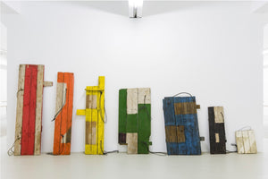 An art installation in a white gallery featuring a row of seven sculptures made from reclaimed wood planks. The sculptures decrease in height from left to right and are painted in different colors: red, orange, yellow, green, blue, black, and white, each with a black cord dangling from it.