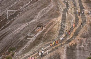 A high-angle photograph shows numerous people climbing a long, winding staircase built into the side of a massive, wet rock hill. Overlaid on the vast rock face are two white text boxes. The upper box asks, Do you spend too much time on social media? The lower box asks, Do you feel well informed by the local news channels?
