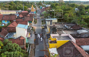 An aerial view of a street in a village in India, lined with colorful houses with red-tiled roofs and surrounded by lush greenery. Several people, some in white robes, walk down the paved road. The image has two text boxes with questions overlaid: Are you sometimes envious of, someone else's achievements? and Have you let someone down lately?
