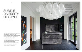 A magazine spread featuring a luxurious kitchen with dark floors, a grey marble island, and black cabinets. A large, cloud-like chandelier made of frosted glass globes hangs over the island. The left page contains text with the headline, Subtle Diversity of Style.