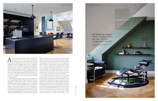 A two-page magazine spread showcasing modern interior design. The left page features a sleek kitchen with matte black cabinetry and an island. The right page shows a reading nook with a green accent wall, a black leather and chrome chaise lounge on a circular rug, and floating shelves.