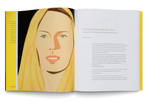 An open art book with a yellow spine laid flat on a white surface. The left page features a large, stylized portrait of a woman with blonde hair and light blue eyes. The right page contains black text on a white background, with the title A Conversation with Alex Katz.