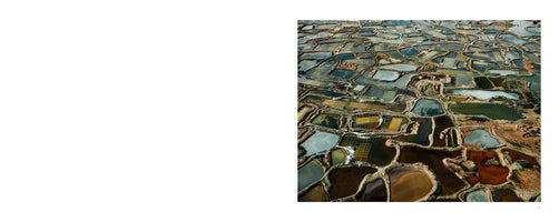 A two-page book spread with a blank white page on the left and a high-angle, aerial photograph of a salt marsh on the right. The photo shows a vast, abstract patchwork of irregularly shaped evaporation ponds separated by earthen dikes. The water in the ponds is a variety of earthy colors, including shades of green, brown, yellow, grey, and rust.