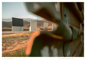 A photograph taken from a close-up, blurry perspective, looking across a field of dry, golden grass toward several prototypes for a border wall. The different wall sections, including one with black vertical slats and others made of solid concrete, stand in a row along a road in a desert landscape with mountains visible in the distance under a pale sky.
