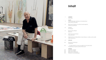 A two-page spread from an art book. On the left, a full-page color photograph shows an older male artist sitting on a bench in his studio. He wears a black polo shirt and paint-stained white pants. Behind him, several large abstract paintings, some featuring upside-down figures, lean against the wall. The right page shows the table of contents in German, with the title Inhalt.