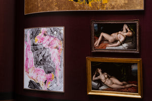 An interior view of an art gallery with a dark red, curved wall. On the left is a large, framed modern abstract painting with bold pink, black, and grey brushstrokes. To the right, two smaller, framed classical paintings of reclining female nudes are stacked vertically. The bottom edge of a large, gold-leafed painting is visible at the top of the frame.