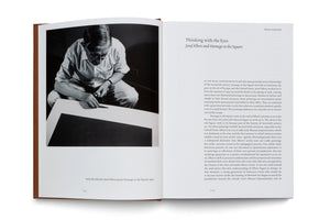 An open art book displays a two-page spread. The left page features a black-and-white photograph of artist Josef Albers, who is crouching and painting his work Homage to the Square in 1950. The right page contains an essay with the title Thinking with the Eyes, Josef Albers and Homage to the Square.