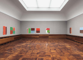 An interior view of a spacious, empty art gallery with a geometric patterned wood floor and a large skylight. Several unframed abstract paintings featuring blocks of red, green, brown, and blue are displayed on the light grey walls above low wood wainscoting.