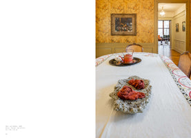 A two-page book spread. The left page is white with a caption. The right page shows a photograph of a dining room with a modern sculpture placed on a white-clothed table. The sculpture is a rough, grey concrete mass topped with two rolled-up red socks resembling flowers. Behind it, a red teapot sits on a tray. The room has yellow floral wallpaper and a view into an adjacent room through a doorway.