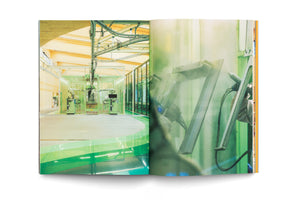 An open book with a two-page photo spread of a modern industrial painting facility. The left page shows a wide shot of a clean factory with robotic arms inside a large, green-framed glass booth. The right page is a hazy, close-up of a gloved worker using a spray gun on a metal frame.