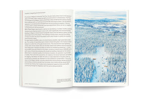 An open book showing a page of text on the left titled Location: Integrating the Environment. The right page is a full-page, aerial photograph of a vast, snow-covered pine forest with a clearing for a construction site, where several vehicles are parked.