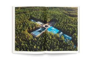An open book displays a two-page aerial photograph of a large, modern, cross-shaped building under construction, nestled within a vast, dense pine forest. The warm, low sunlight illuminates the scene, highlighting the treetops.
