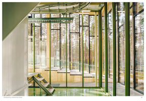 An interior view of a modern building featuring a glossy green floor and industrial metal steps. Floor-to-ceiling glass walls, framed with green metal and light wood beams, offer a clear view of a dense pine forest outside. An overhead conveyor track is visible near the ceiling.