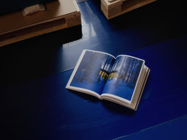 An open book lies on a glossy, deep blue surface, showing a two-page photograph of a modern, illuminated building nestled in a snowy forest at dusk. Wooden pallets are visible in the background.