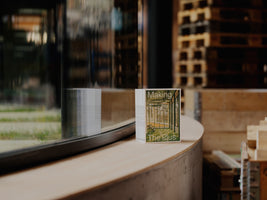 A close-up of a small booklet titled Making The Plus standing on a curved wooden windowsill. The booklet's cover features a green-hued architectural image. The window reflects the booklet, and the out-of-focus background shows an industrial space with stacked wooden pallets.