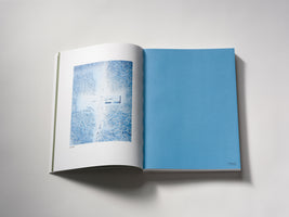 A high-angle photograph of an open book on a plain white surface. The left page displays an aerial, blue-toned image of buildings in a snow-covered forest. The right page is a solid, uniform light blue.
