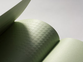 A minimalist close-up of an open notebook with sage green pages. One page is embossed with a tactile pattern of small circles, with a distinct shadow falling diagonally across it, highlighting the texture against a plain background.