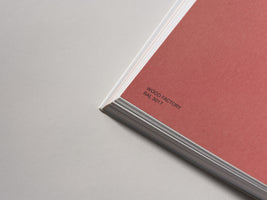 A close-up, angled shot of the corner of a stack of paper on a light grey surface. The top page is a textured, dusty rose color with the words WOOD FACTORY and RAL 3017 printed in black. The white edges of the pages below are visible.