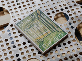 An overhead, angled shot of a book titled Making The Plus resting on a sheet of perforated silver metal. The book's cover features a green-toned architectural illustration with a textured, bumpy surface. Below the metal sheet, a wooden surface is visible through the circular holes.