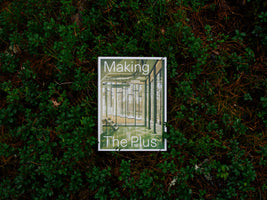 A top-down photograph of a book titled Making The Plus resting on a bed of dark green forest foliage and brown pine needles. The book's cover shows the interior of a modern, glass-walled building, contrasting with its natural surroundings in the photo.