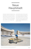 A magazine page in German titled Neue Hauptstadt. A large photo shows a construction worker in a blue shirt shoveling sand on a vast, dusty plaza. In the background stands a monumental, light-colored building with a massive, curved colonnade under a clear blue sky. Other workers are visible in the distance.