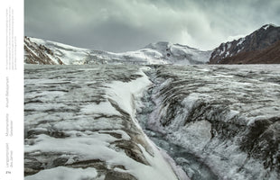 A stark, wide-angle view from the surface of a massive glacier, where a meltwater stream carves a channel through the dirty ice and snow in the foreground. In the distance, towering snow-covered mountains rise against a heavy, overcast sky.