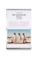 An overhead shot of an open book on a white background. The page is in German, with the headline Der sterbende Fluss, and features a photograph of three beekeepers in full white protective suits standing in a desert landscape in front of beehives, surrounded by a swarm of bees.