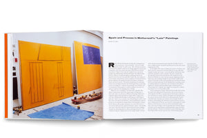 A two-page spread from an art book. The left page is a full-page color photograph of Robert Motherwell's studio, showing large abstract canvases in yellow-orange and blue leaning against a curved wall. The studio floor is paint-splattered and holds a blue cloth and a low cart with paint jars. The right page features the title and beginning of an essay, Spain and Process in Motherwell's Late Paintings.