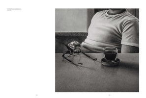 A two-page book spread, with a realistic, grainy black and white drawing on the right page. The drawing shows a person in a t-shirt sitting at a table. In the foreground, a pair of eyeglasses and a double-walled glass with a dark drink rest on the tabletop.