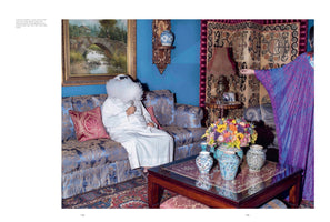 A man in a white thobe and headdress sits on an ornate blue sofa, his face completely hidden by a massive cloud of white vapor from a vape pen he is holding. He is in a lavishly decorated room with bright blue walls, a landscape painting, patterned rugs, and a coffee table with a large, colorful bouquet of flowers. To the right, another person in a purple and blue robe is partially visible.