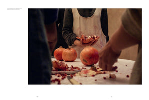 A close-up photograph shows several people gathered around a table preparing pomegranates. The person in the center wears a white apron stained with red juice and holds half of a pomegranate that has been broken open. The table is covered with whole pomegranates, scattered red seeds, and pieces of rind.