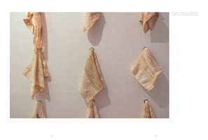 A photograph in a book shows an art installation on a white wall. Several pieces of pale, peach-colored fabric with different textures, including lace and waffle weave, hang from small rings in a scattered grid pattern, casting soft shadows.