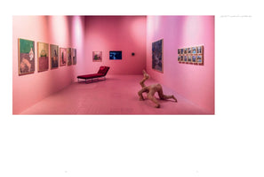 An interior view of a modern art gallery with bright pink walls and a pink tiled floor. The room features various paintings, a red chaise lounge, and a sculpture of a headless, armless human lower body crawling on the ground.