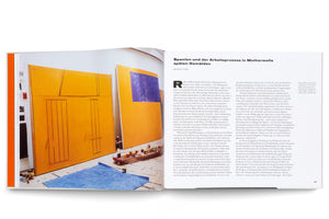 A two-page spread from an art book about Robert Motherwell. The left page shows a photograph of his studio, with two large, yellow abstract paintings leaning against a wall. On the paint-splattered floor are jars of paint on a low rolling cart and a piece of blue fabric. The right page features an article in German about the artist's work.