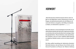 A two-page spread from an art catalogue. On the left, a photograph shows an art installation: a speaker's podium covered in a mosaic of small, silver mirror tiles. A red LED display on the podium reads OUT FEMINISM. A microphone on a stand is positioned next to it. The right page contains the foreword, titled VORWORT, in German.