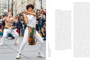 A two-page book spread with text on the right and a photo on the left. The photo shows a Black woman and a shirtless white man dancing energetically on a city street. Both wear white pants, colorful sashes, and have white paint on their faces as a crowd watches and takes pictures.